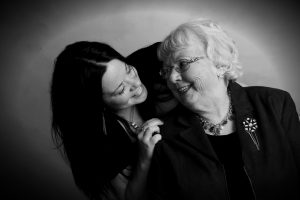 Photo: A young woman cheerfully leans in over the shoulder of a happy elderly woman. They are smiling at each other.